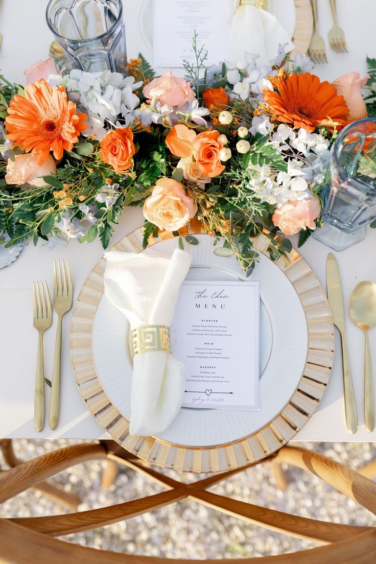 an elegant table setting with orange and white flowers on the centerpiece, gold cutlery, and silverware