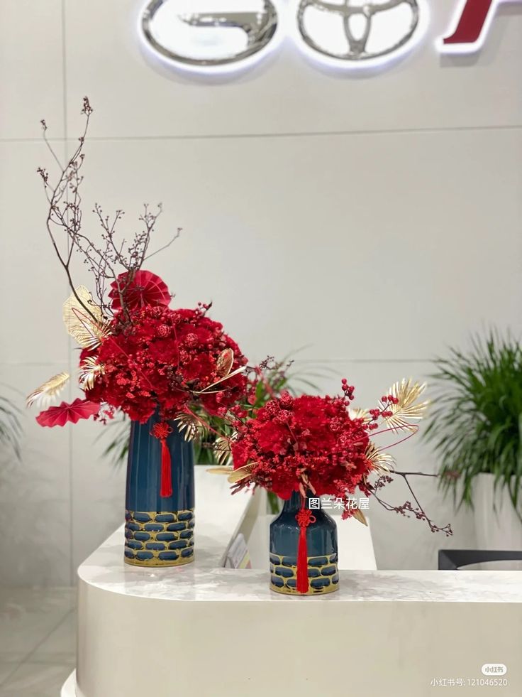 two blue vases with red flowers in them on a white table next to a plant