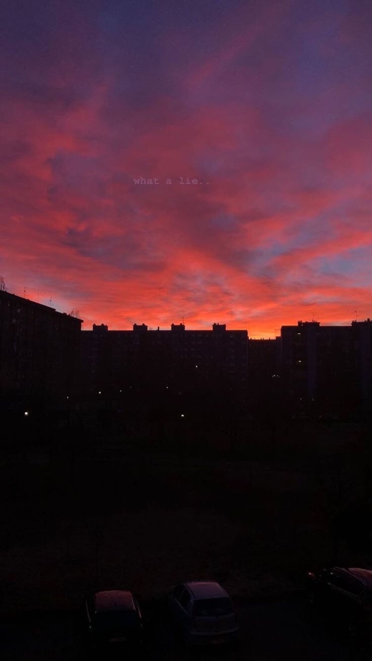 the sun is setting over some buildings and cars parked in a parking lot at night