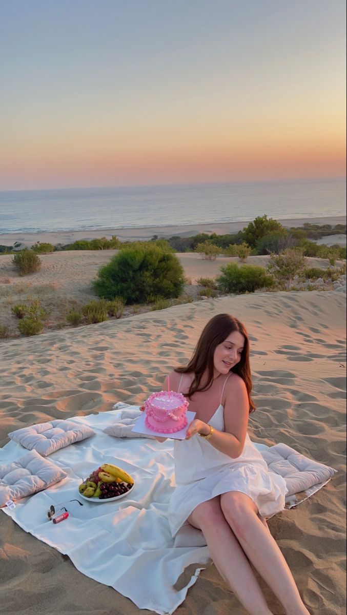 a woman sitting in the sand with a pink cake