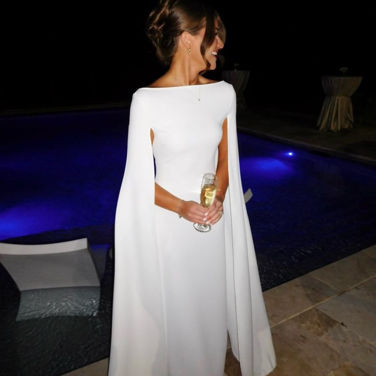 a woman standing next to a pool at night wearing a white dress with cape on it