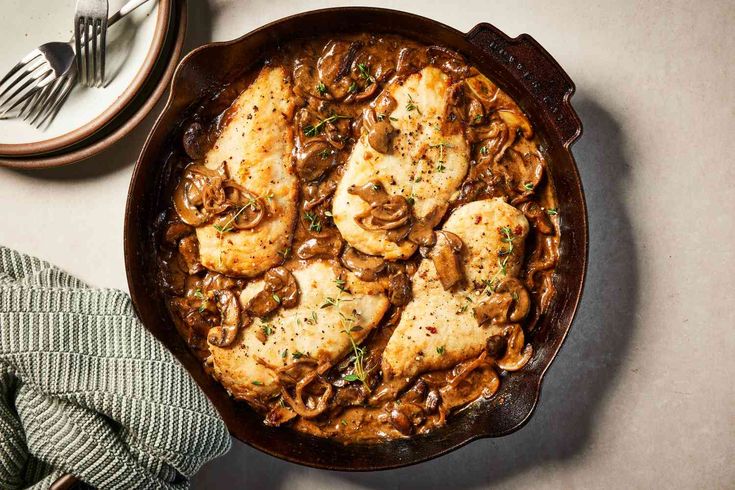 a skillet with chicken, mushrooms and sauce in it next to a plate of silverware