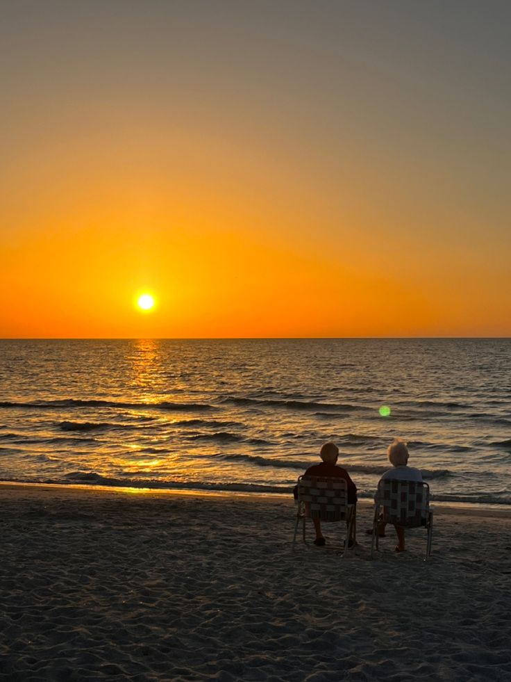 two people are sitting on the beach watching the sun go down over the ocean and water