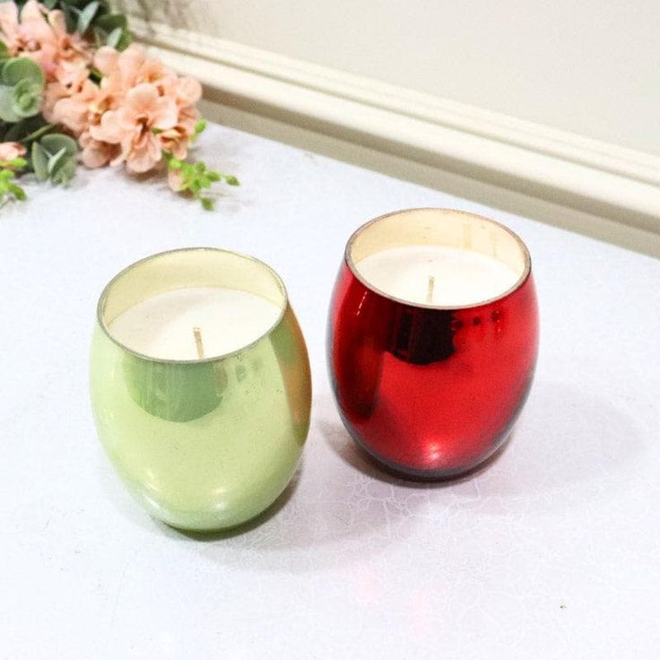 two red and green candles sitting next to each other on a white table with flowers in the background