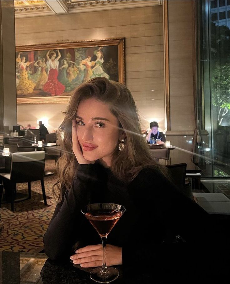 a woman sitting at a table with a wine glass in front of her and looking into the camera