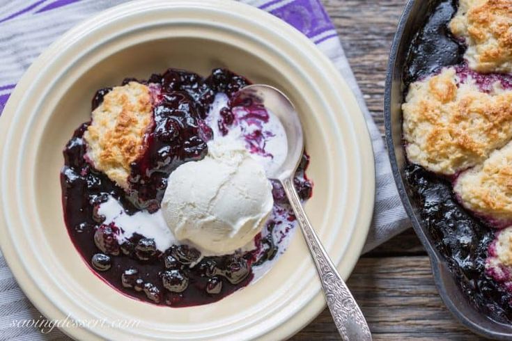 blueberry cobbler with ice cream in a bowl on a wooden table next to a baking dish