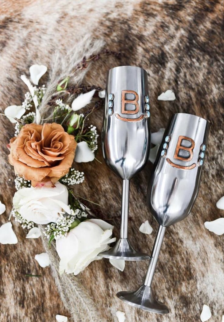 two silver wine glasses sitting on top of a table next to white flowers and petals