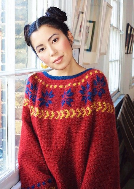 a woman standing in front of a window wearing a red sweater with blue and yellow designs