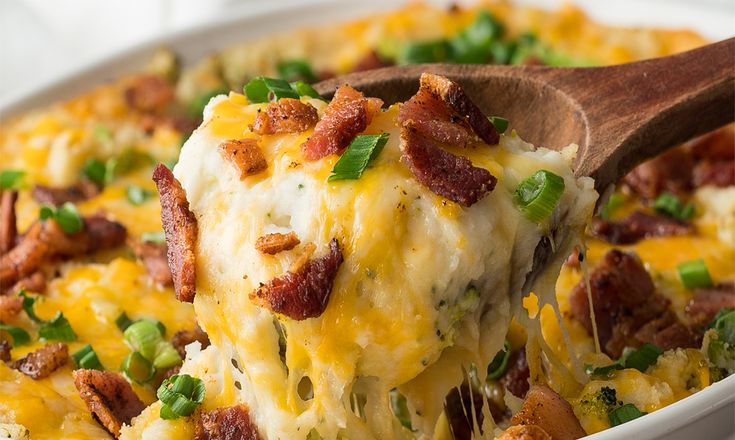 a wooden spoon scooping some food out of a casserole dish with bacon and green onions