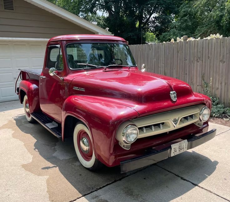 an old red truck is parked in front of a house with a fence behind it