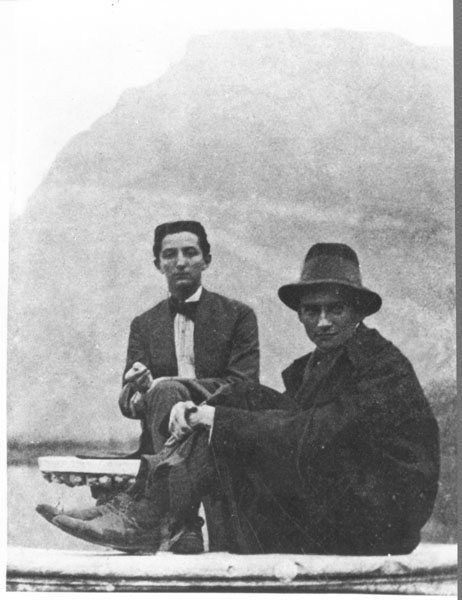 two men in suits and hats sitting next to each other on the ground with mountains in the background