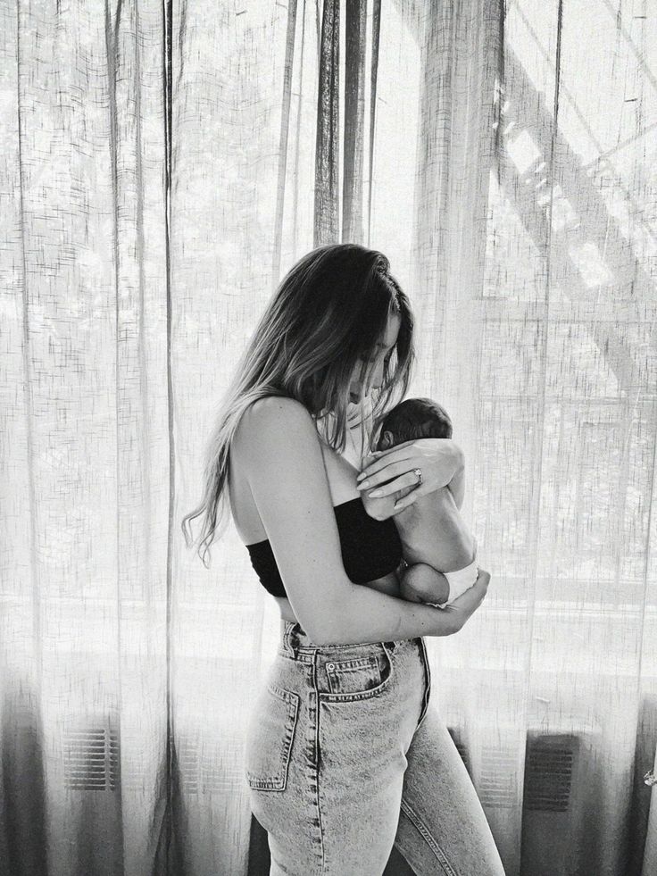 a woman holding a baby in her arms while standing next to a window with curtains