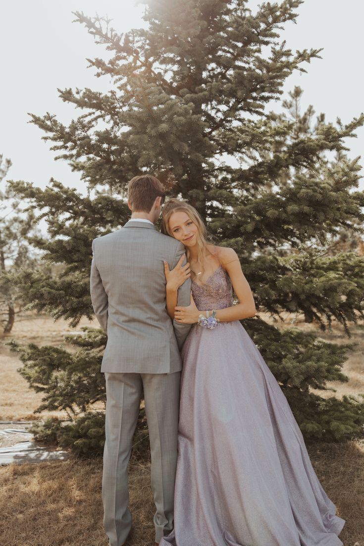 a man and woman standing next to each other in front of a tree with their arms around each other