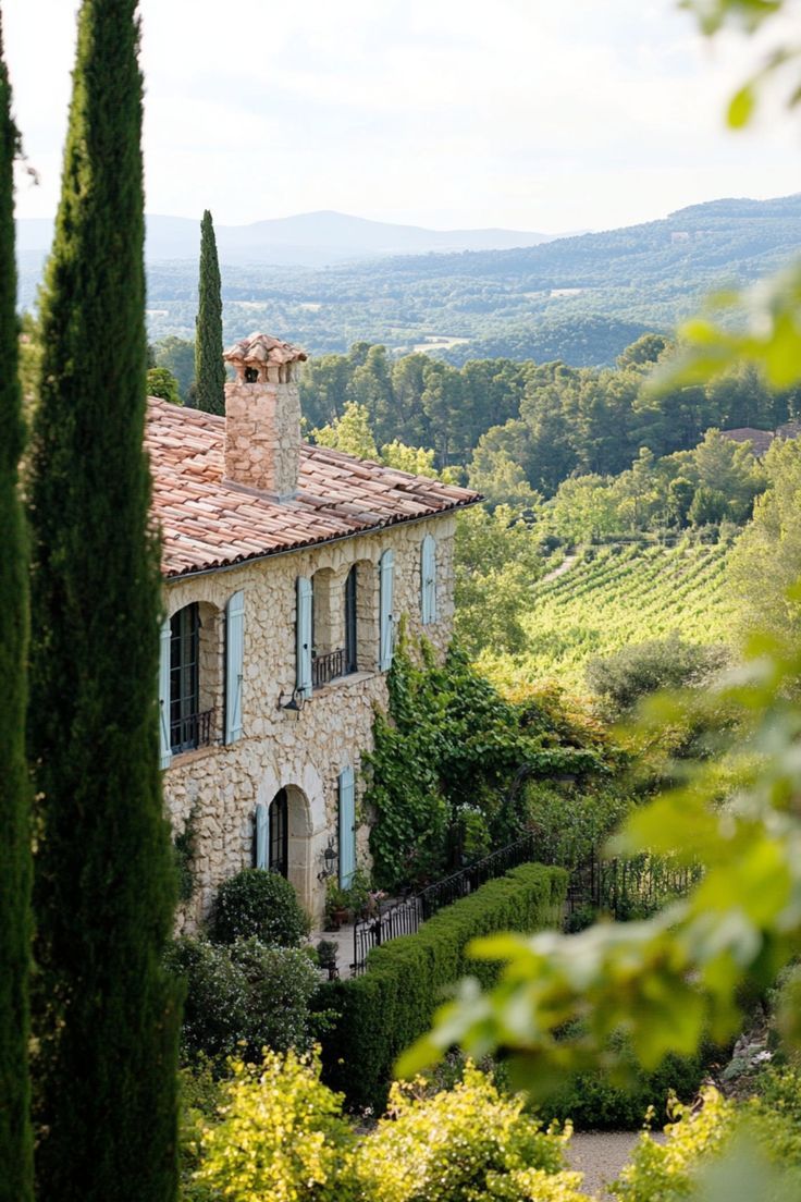 an old stone house surrounded by trees and greenery with mountains in the back ground