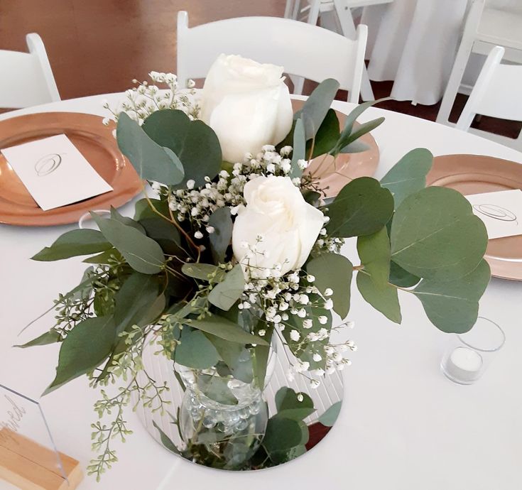 a vase filled with white roses and greenery on top of a dining room table