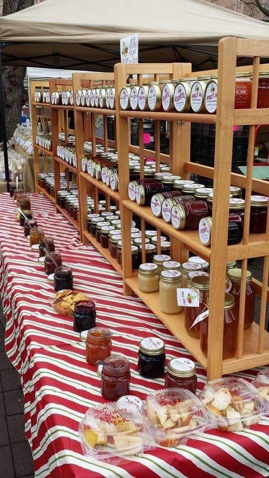 many jars of jams are on display at an outdoor vendor's table with red and white striped cloth