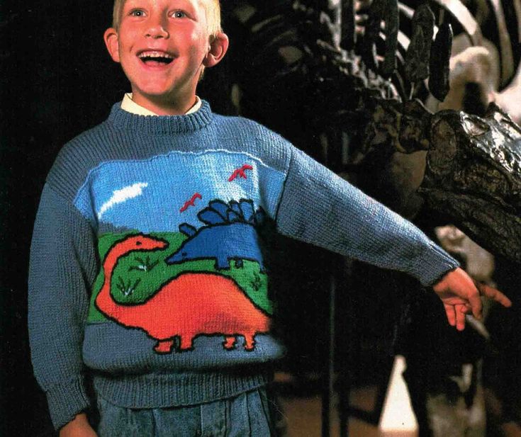 a young boy standing in front of an adult dinosaur statue wearing a sweater with dinosaurs on it