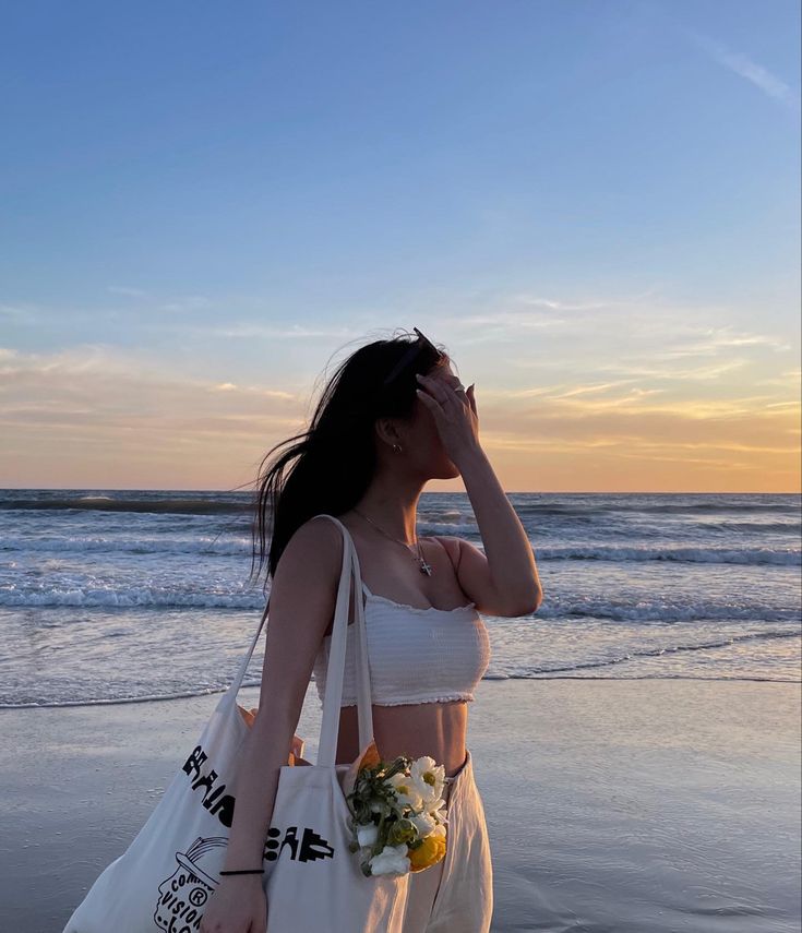 a woman standing on the beach with a bag in her hand and flowers in her other hand