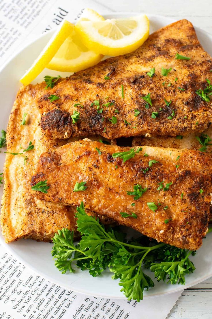 two pieces of fish on a white plate with parsley and lemon wedges next to it