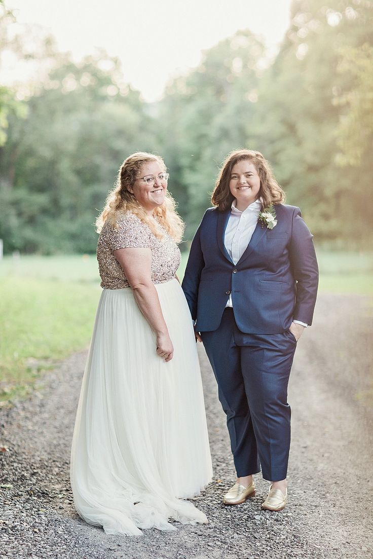 two women standing next to each other on a dirt road in front of some trees