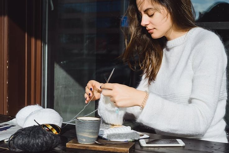 a woman sitting at a table with a cup and spoon in her hand while knitting