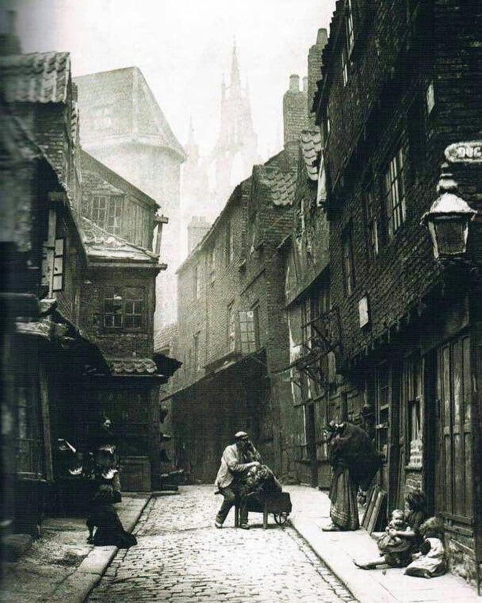 an old black and white photo of people walking down a narrow alleyway with buildings in the background