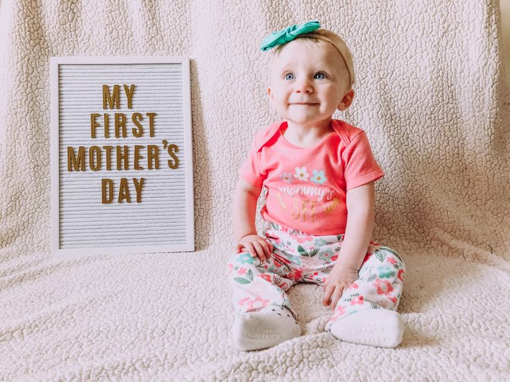 a baby sitting on a bed next to a sign that says my first mother's day