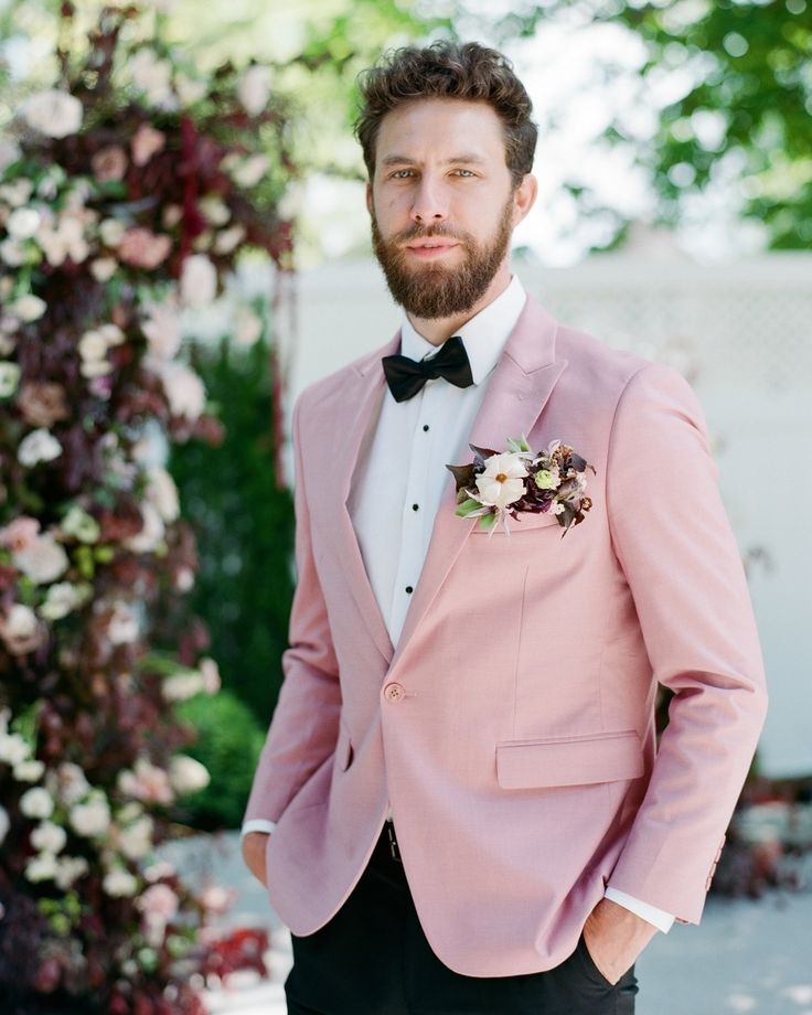 a man wearing a pink suit and bow tie standing in front of a floral arch