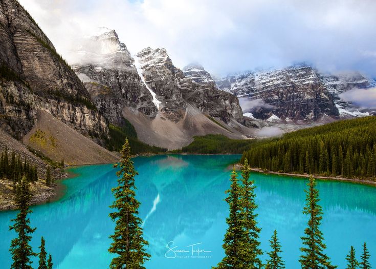 the blue water is surrounded by pine trees and snow capped mountain peaks in the distance