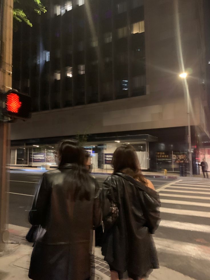 two people walking down the street in front of a tall building at night with red traffic lights