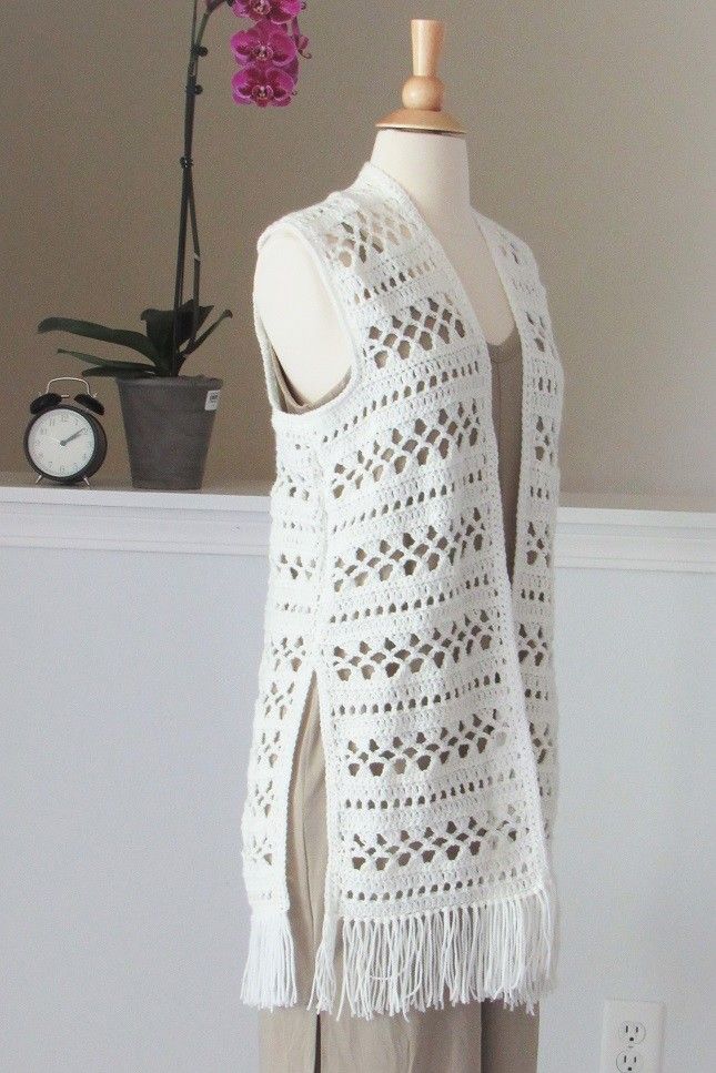 a white crocheted vest with tassels on the sides and a flower pot behind it