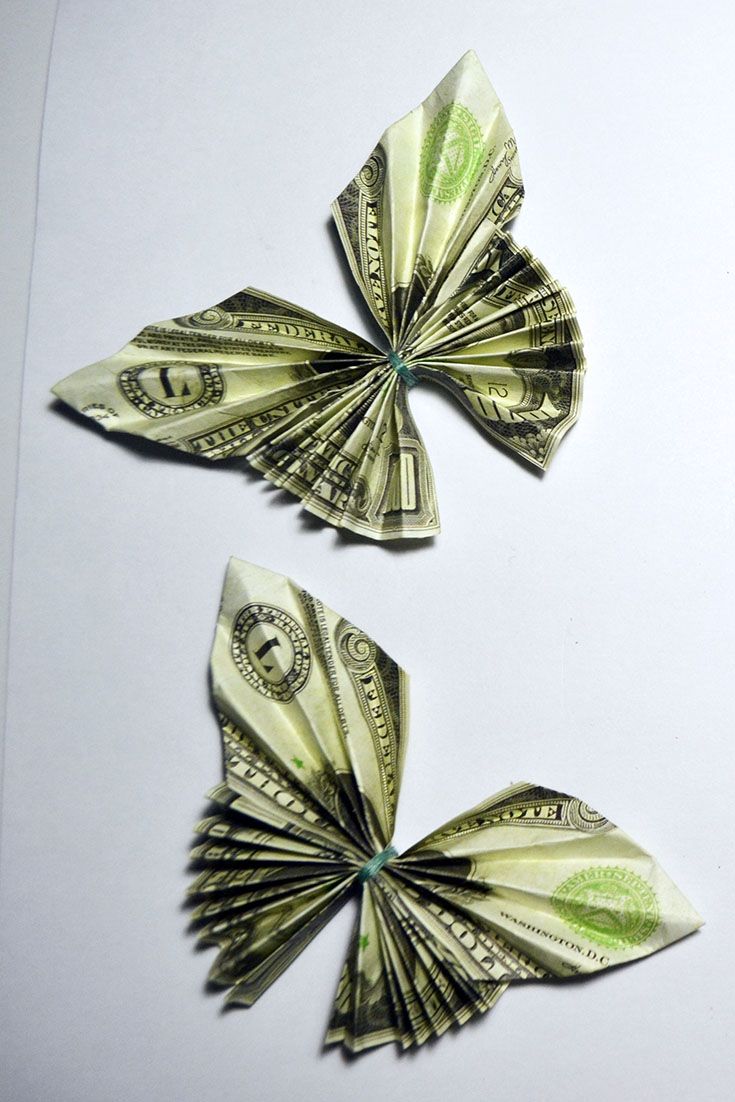 two pieces of paper folded in the shape of butterflies on a white surface with one dollar bill sticking out of it