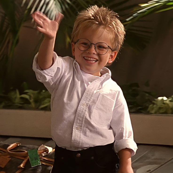 a young boy wearing glasses and holding his hand up in the air while standing next to a plant