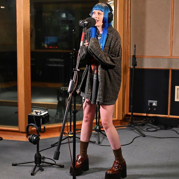 a woman with blue hair is singing into a microphone in front of a recording studio