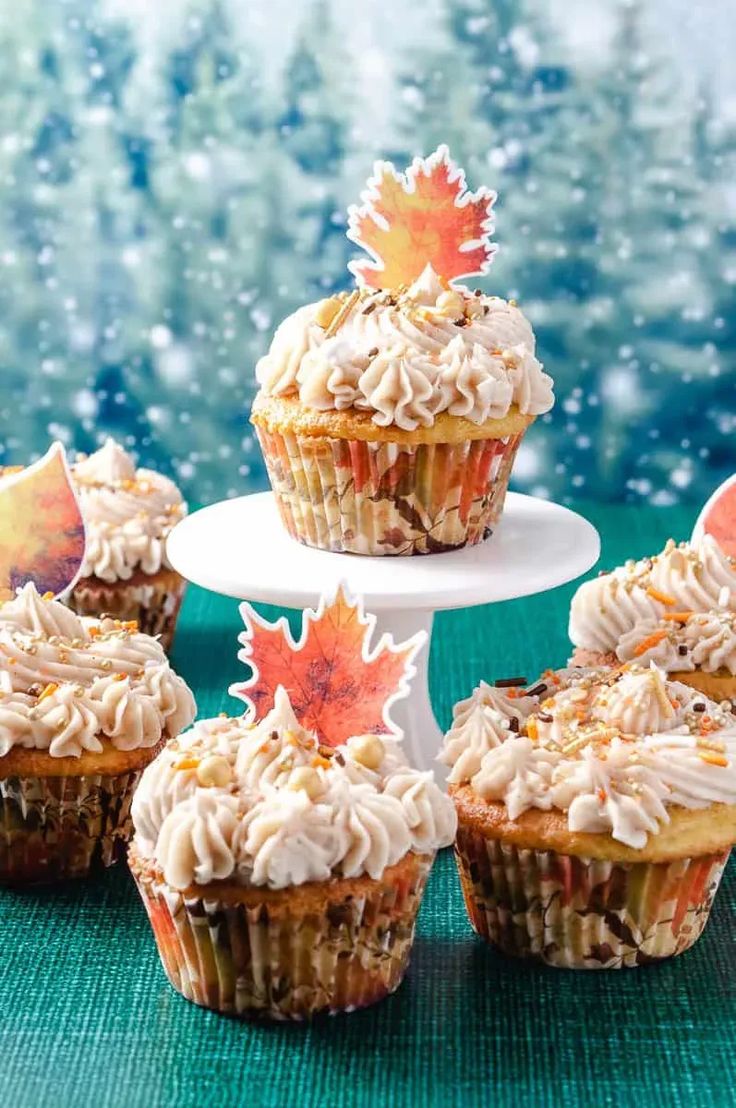 cupcakes with frosting and maple leaf decorations on a green tablecloth in front of snow - covered trees