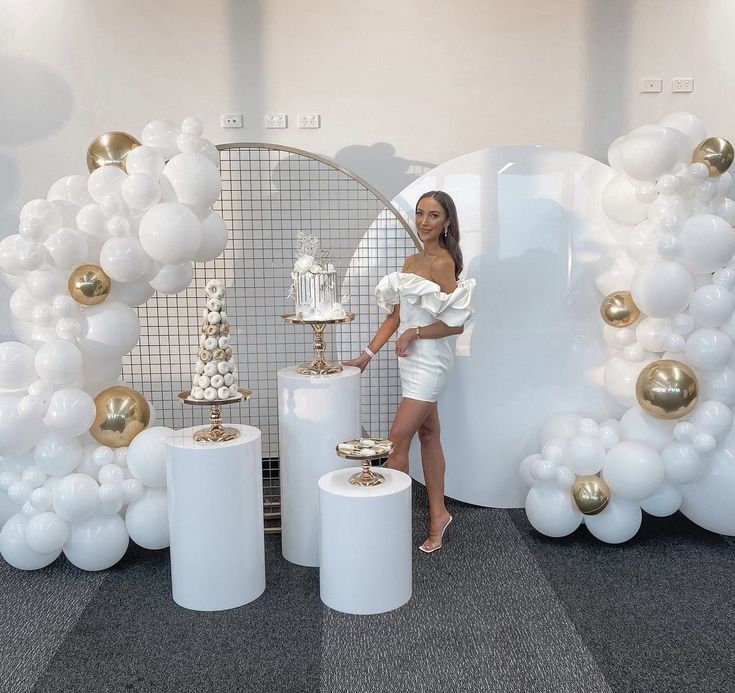 a woman standing in front of balloons and cake on top of white pedestals with gold accents