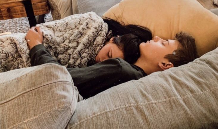 two people laying on top of a couch covered in blankets and pillows with their eyes closed