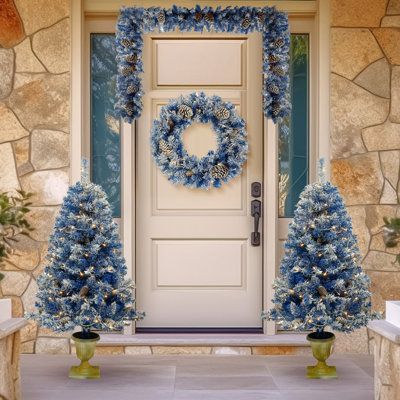 a front door decorated for christmas with blue wreaths and potted trees on either side