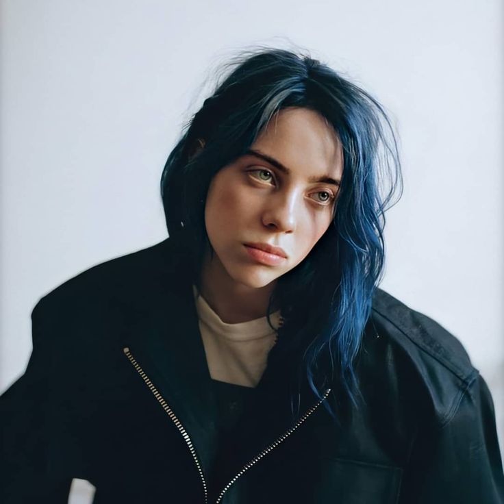 a woman with blue hair wearing a black jacket and looking at the camera while standing in front of a white wall