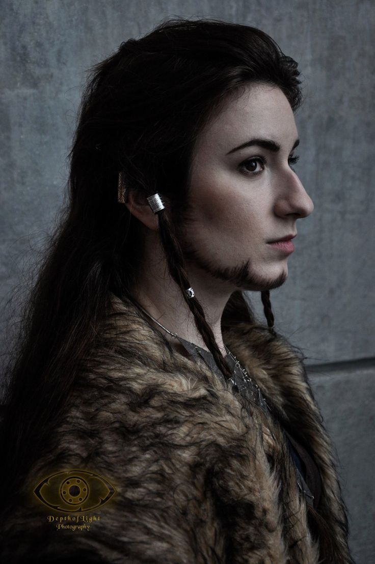 a woman with long hair and piercings on her ear wearing a fur coat against a concrete wall