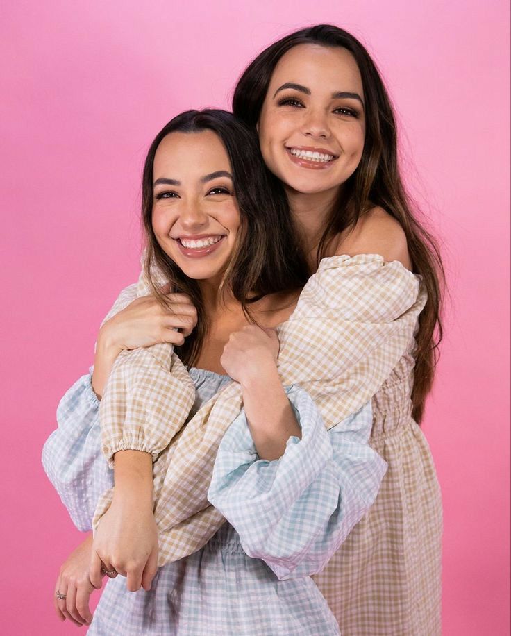 two young women are hugging each other in front of a pink background and one has her arms around the other's shoulders