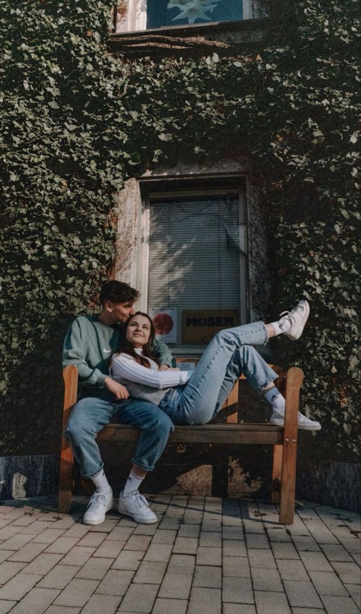 a man and woman sitting on a bench in front of a building with ivy covered walls