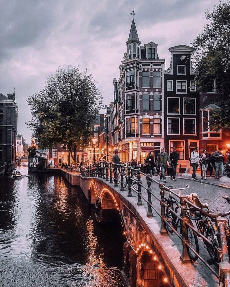 people are walking along the side of a river at night with buildings in the background