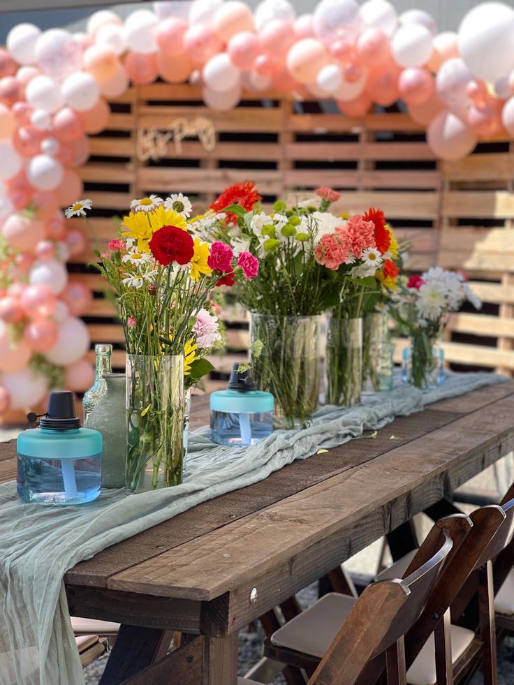 a long table with vases filled with flowers on top of it and balloons in the background