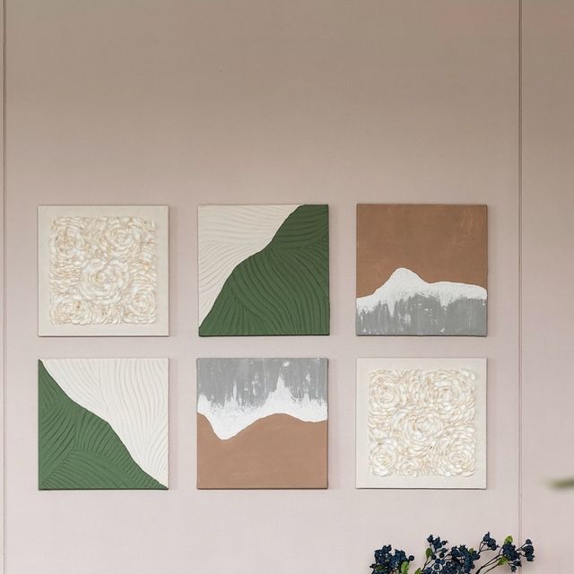 four square paintings hang on the wall above a vase with blue flowers in front of it