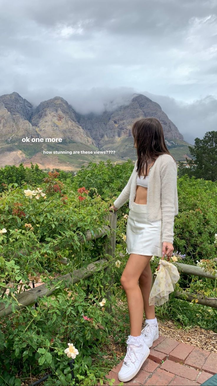 a woman standing on top of a lush green hillside next to flowers and trees with mountains in the background