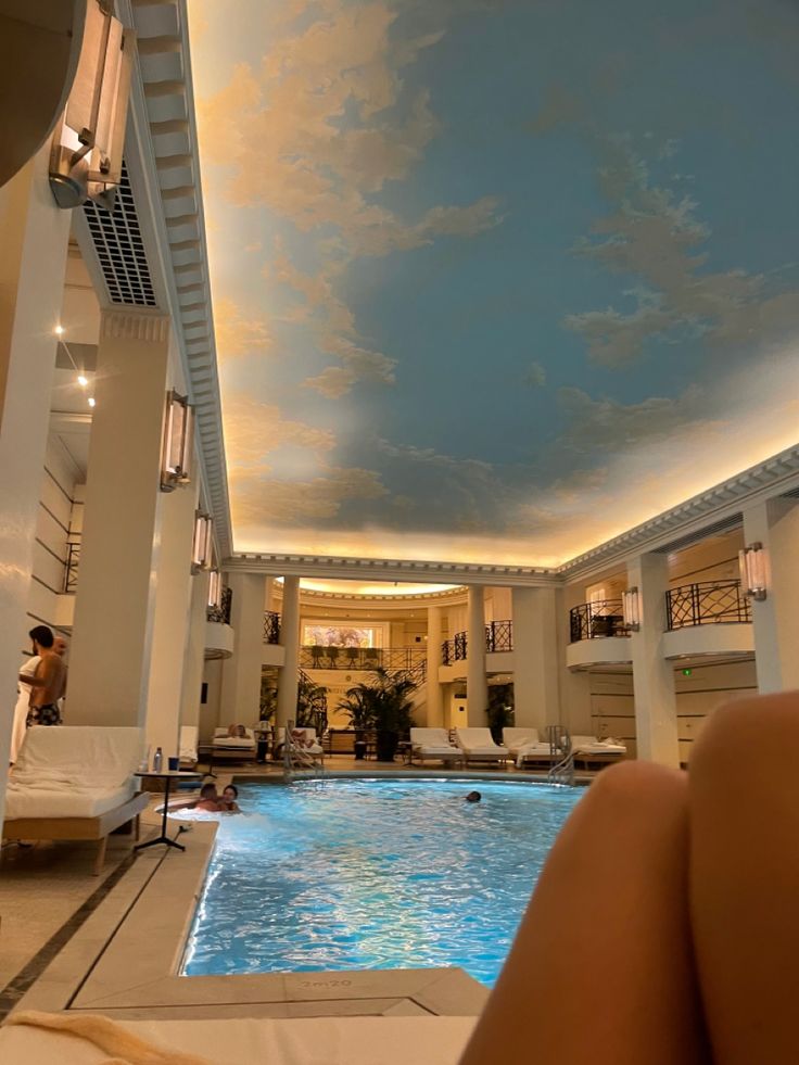 an indoor swimming pool with sky painted on the ceiling and people lounging in lounge chairs