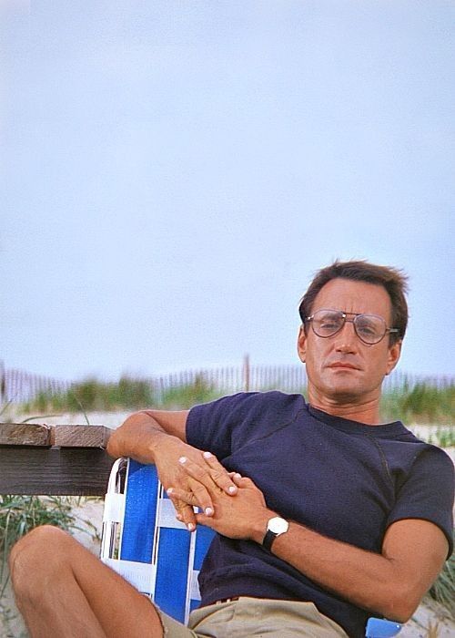 a man sitting in a chair on the beach with his arm crossed and looking off into the distance