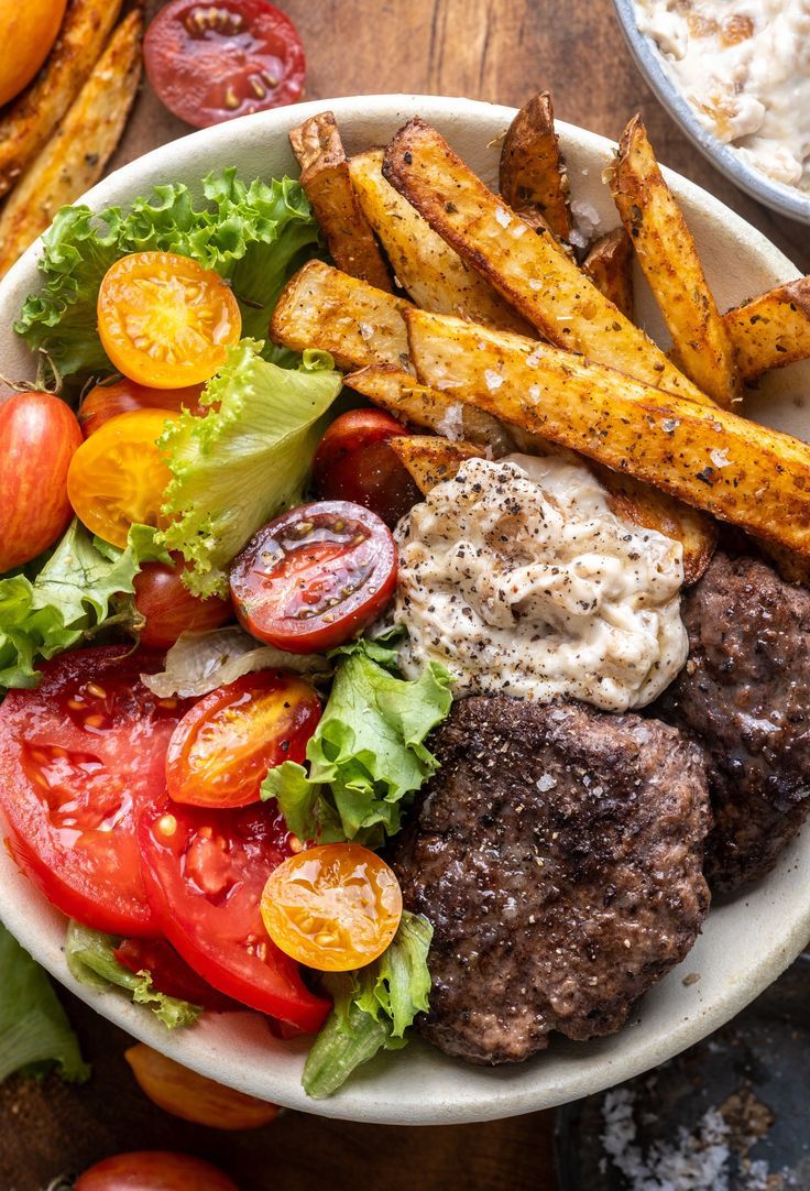 a white bowl filled with meat and vegetables next to some fries on a wooden table