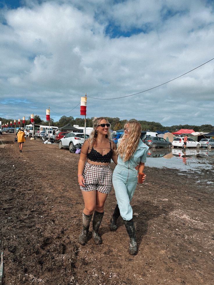 two women in short shorts and cowboy boots are walking on the beach with boats behind them